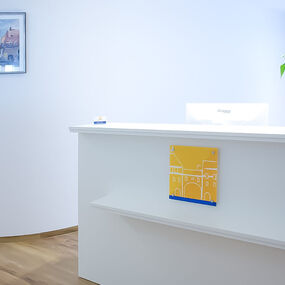 Heller Empfangsbereich einer modernen Arztpraxis mit Pflanze und Computer am Tresen.